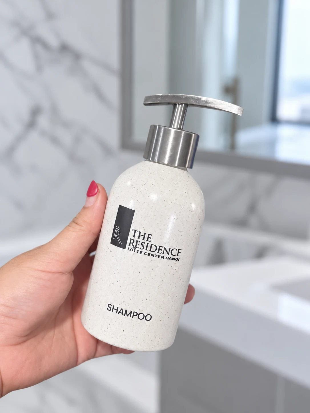 Bình trụ tròn 180 ml vòi inox với chữ nhận diện trên thân, được cầm nghiêng trong không gian sáng