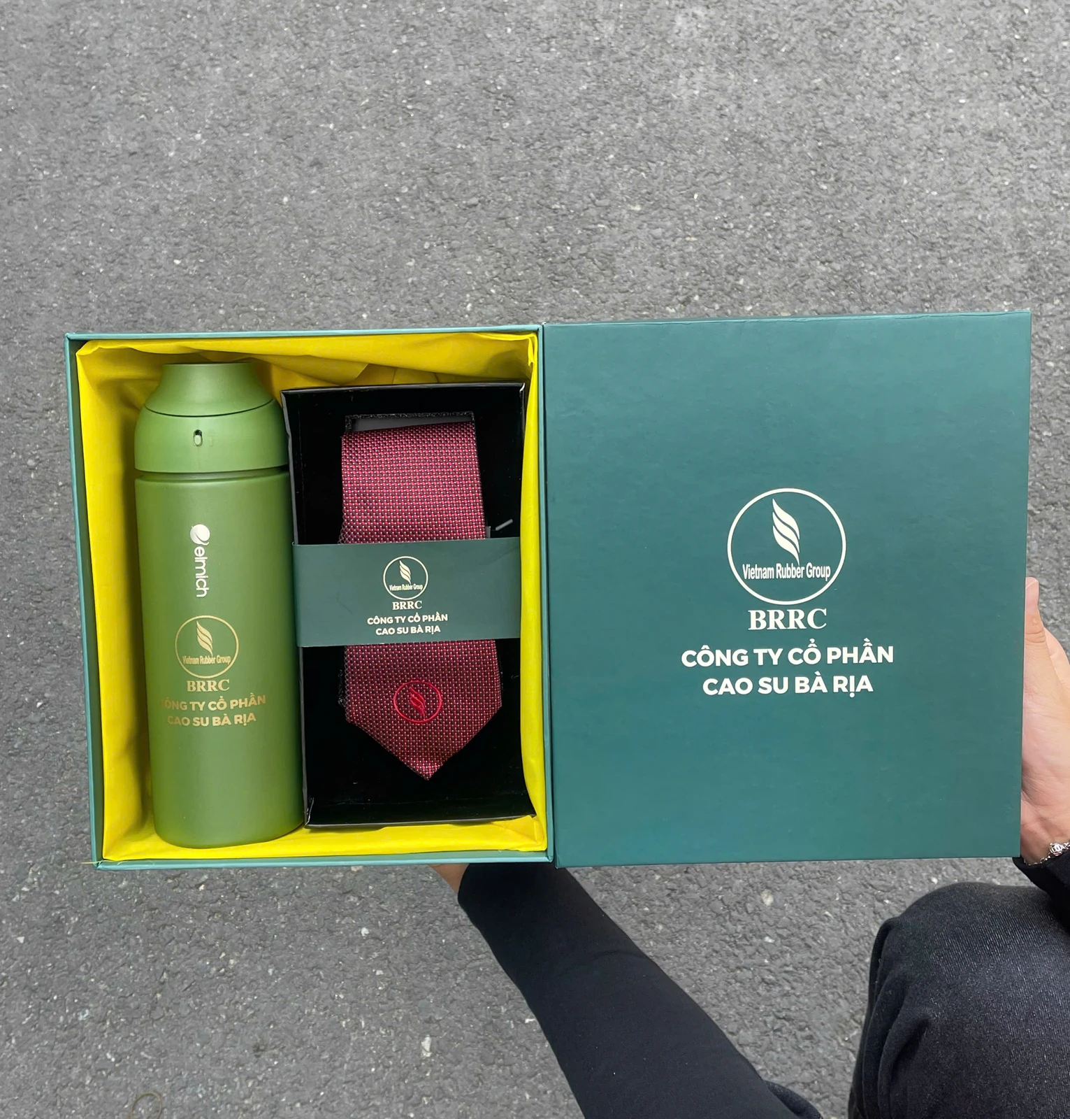 Hộp xanh mở có bình và cà vạt, cạnh nắp hộp mang nhận diện BRRC