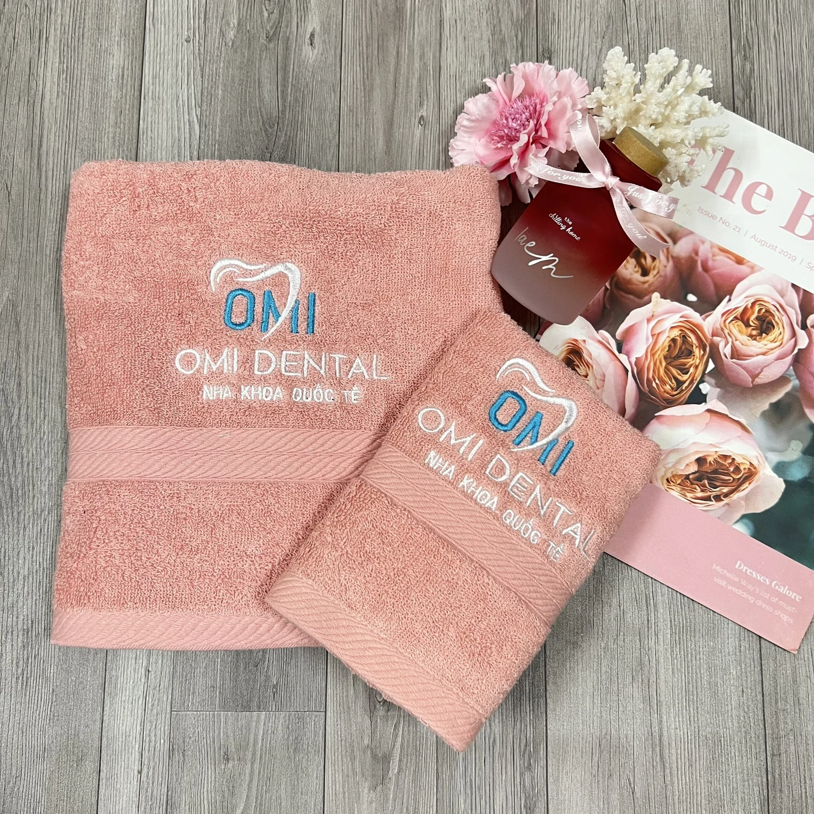 Hai khăn màu hồng với logo OMI Dental thêu xanh trắng