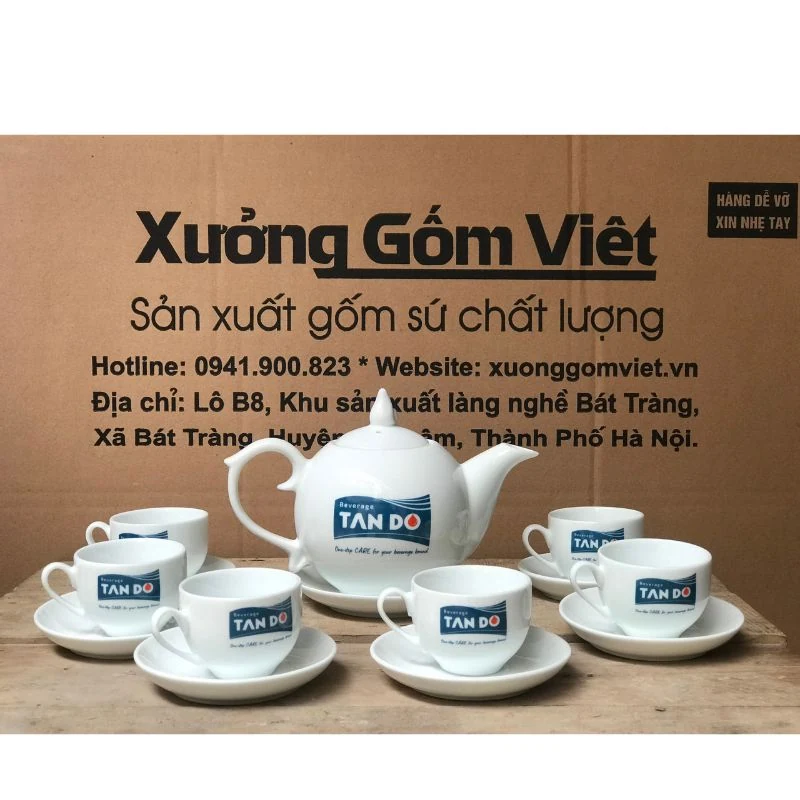 Logo xanh trên ấm và chén trắng; ảnh có thêm đĩa kê ấm.