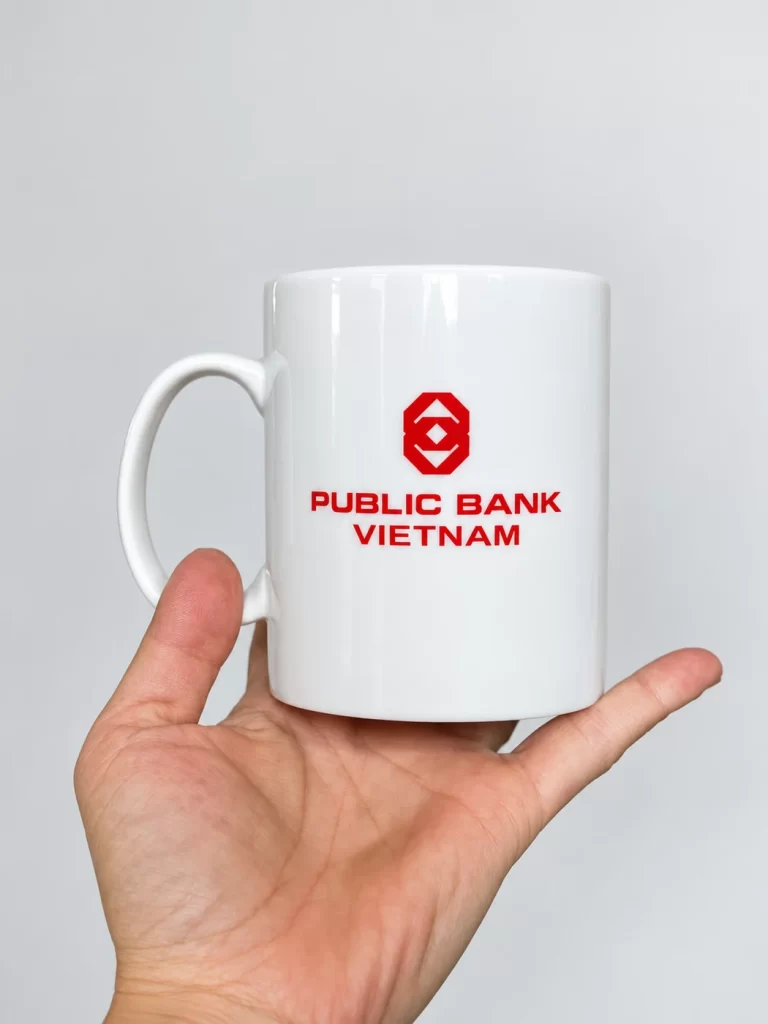 Logo đỏ trên mặt ly sứ trắng, quai ở bên trái và bàn tay nâng ly