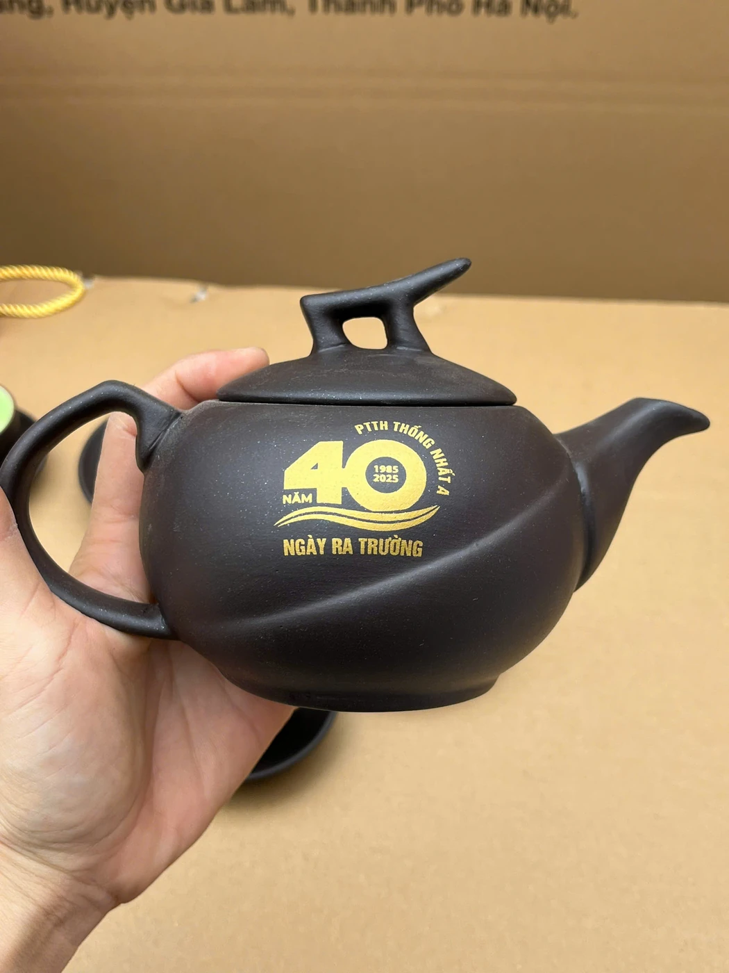 Ảnh thật thân ấm đen có chữ kỷ niệm màu vàng sáng và bề mặt cong khi cầm