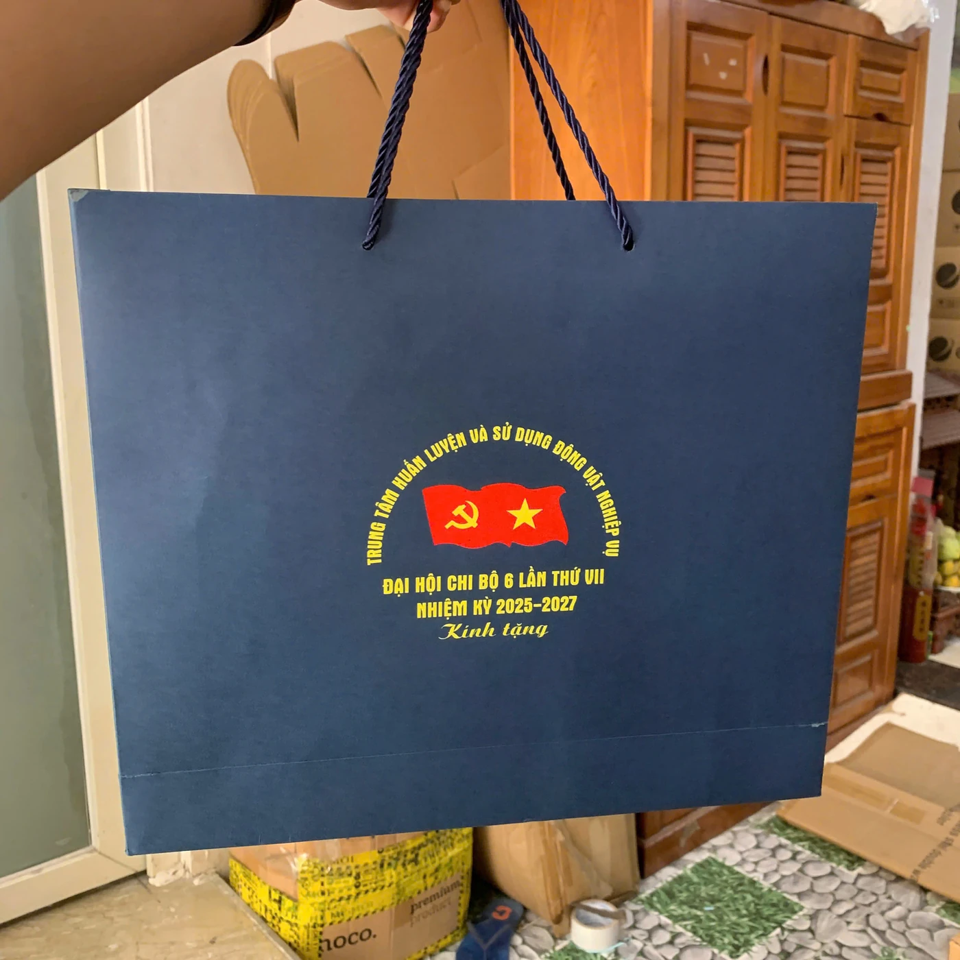 Ảnh thật túi giấy xanh được cầm bằng quai dây, mặt túi có đường gấp gần đáy và nhận diện chương trình
