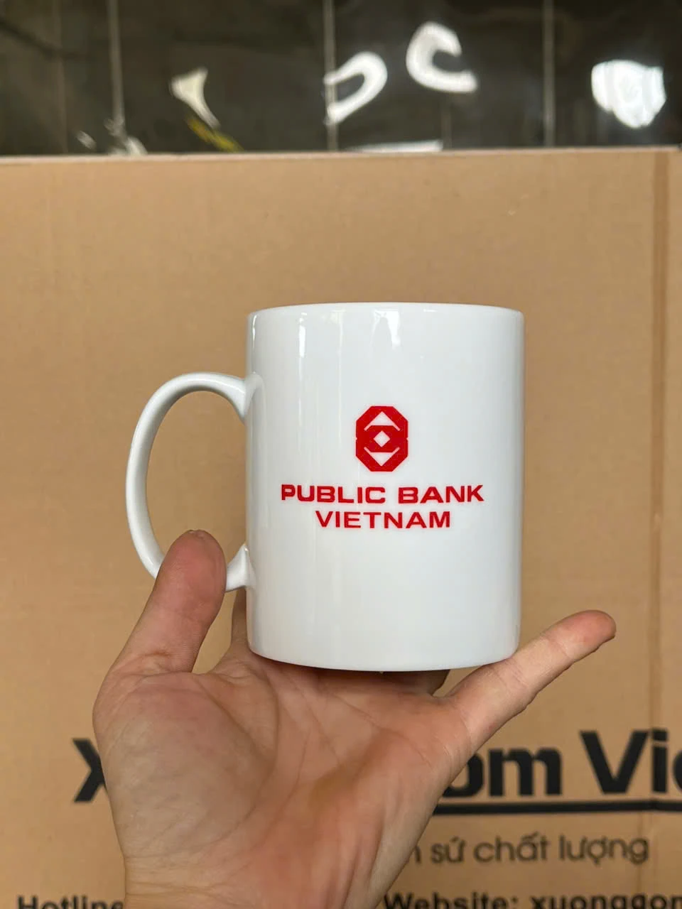 Ảnh thật cốc trắng cầm trên tay, quai bên trái và logo PUBLIC BANK VIETNAM quay về phía người xem