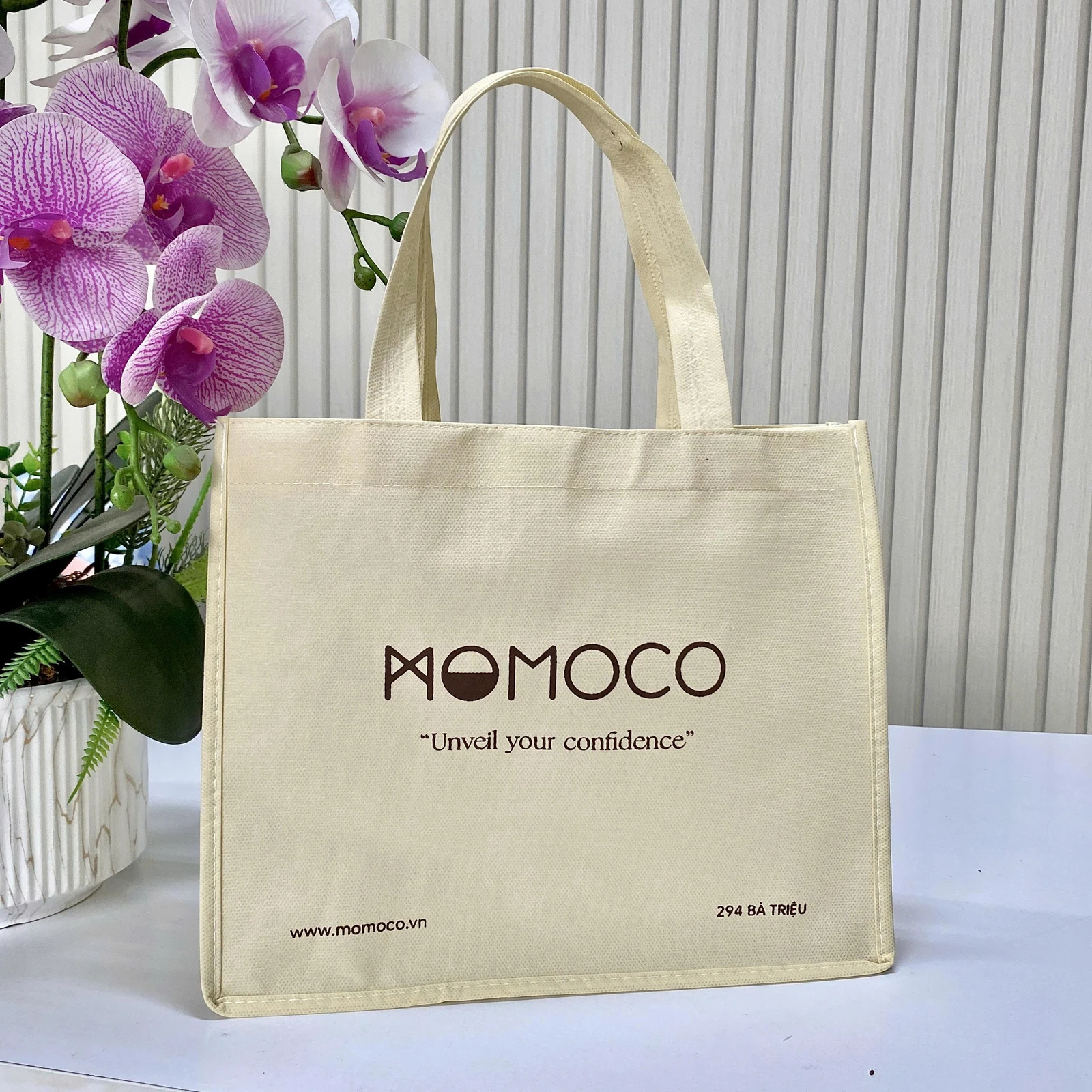 Ảnh nguồn túi màu kem có hai quai dẹt, nhận diện MOMOCO ở mặt trước