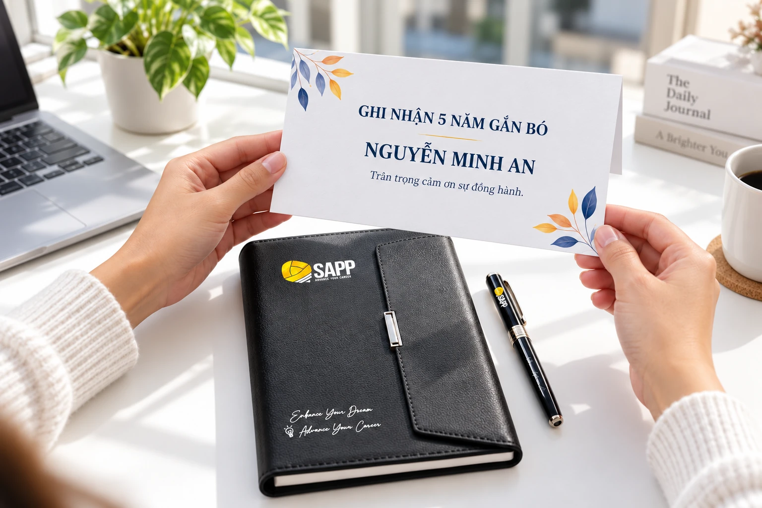 Thiệp ghi nhận 5 năm và tên giả định cầm trên bộ sổ, bút có nhận diện SAPP