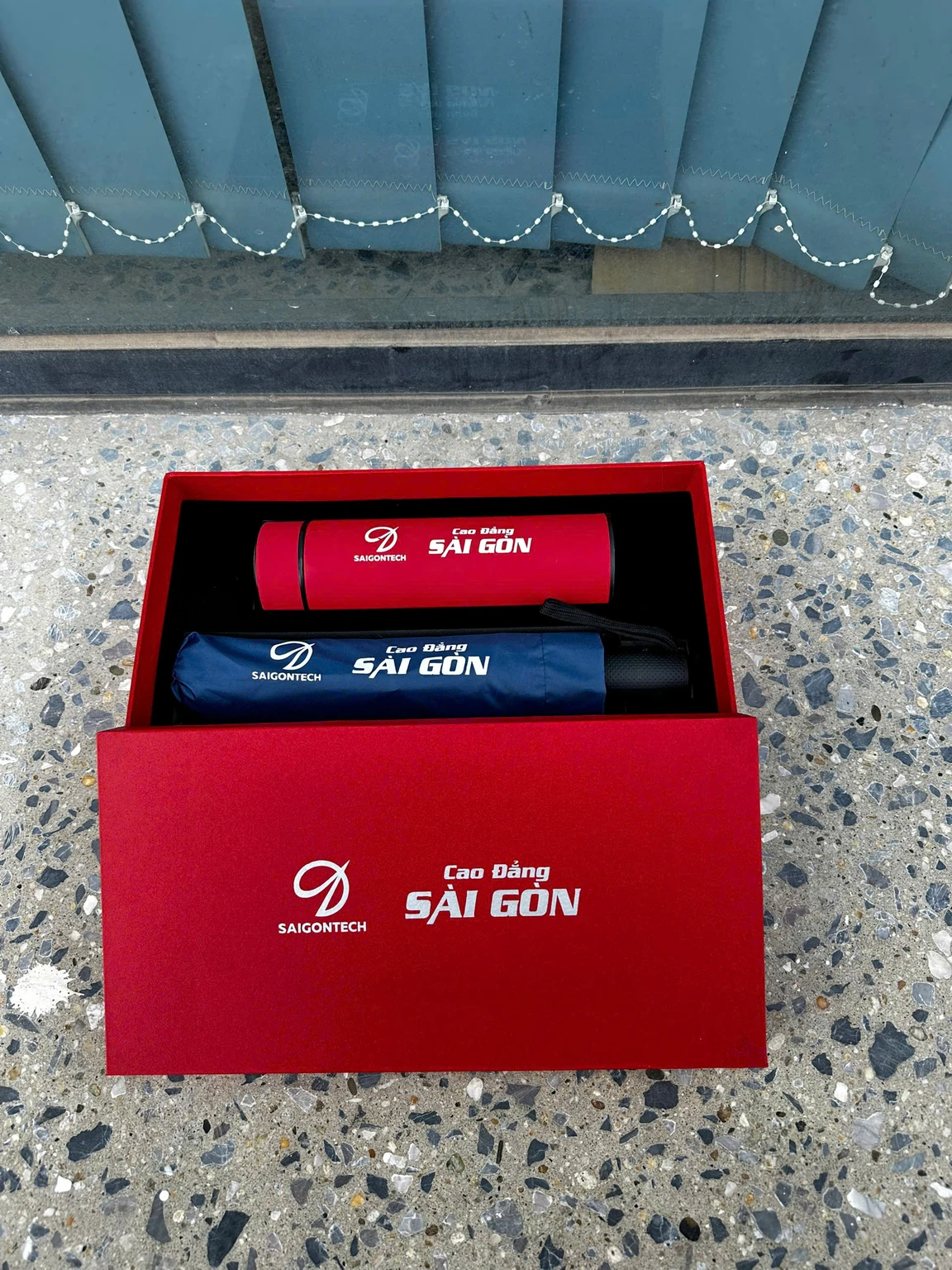 Ảnh thật logo SAIGONTECH và Cao Đẳng Sài Gòn trên bình đỏ, dù xanh và hộp đỏ